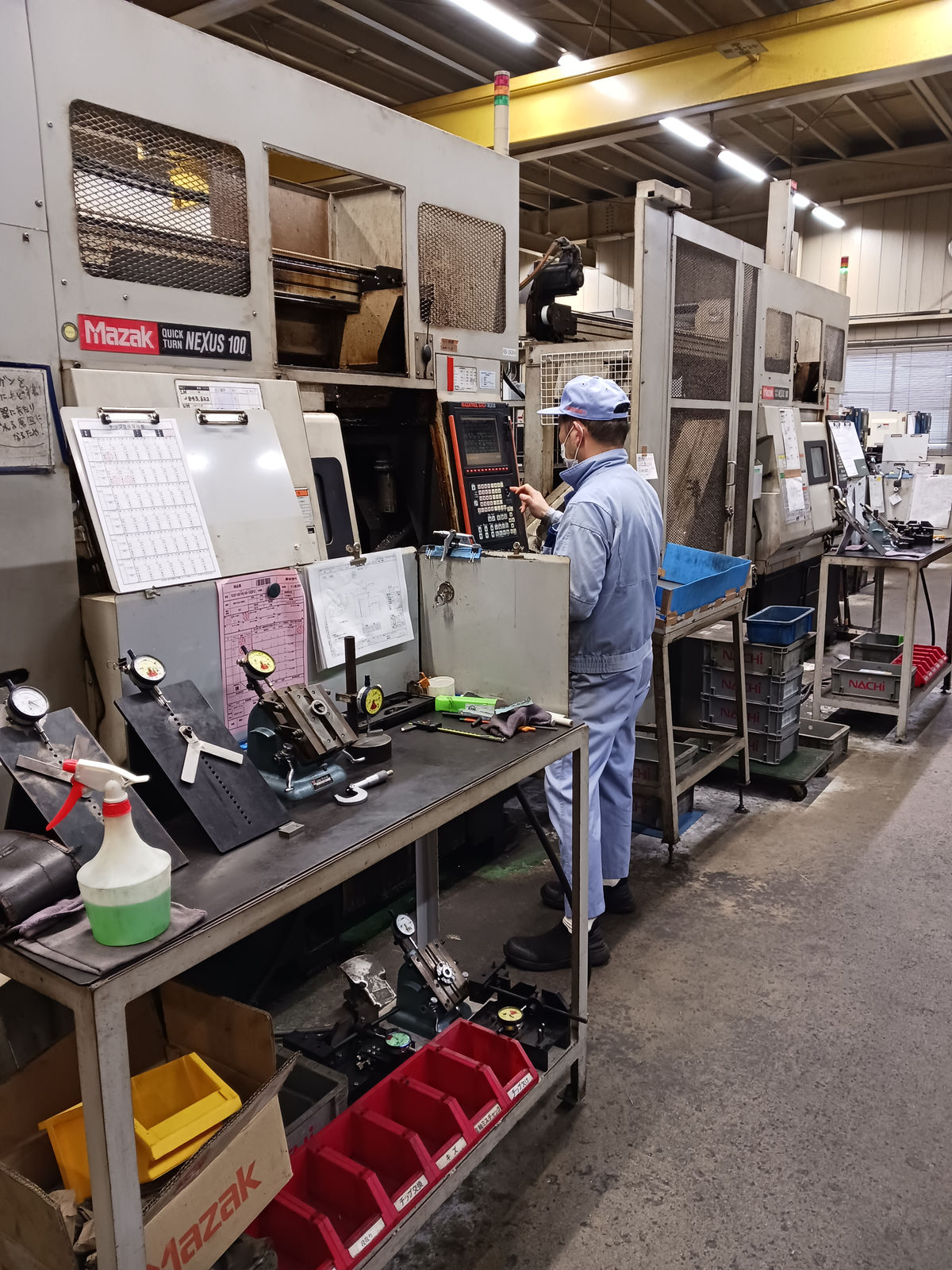 マザック QUICK TURN NEXUS 100の前で操作盤に触れる作業者と、測定器を並べた作業台